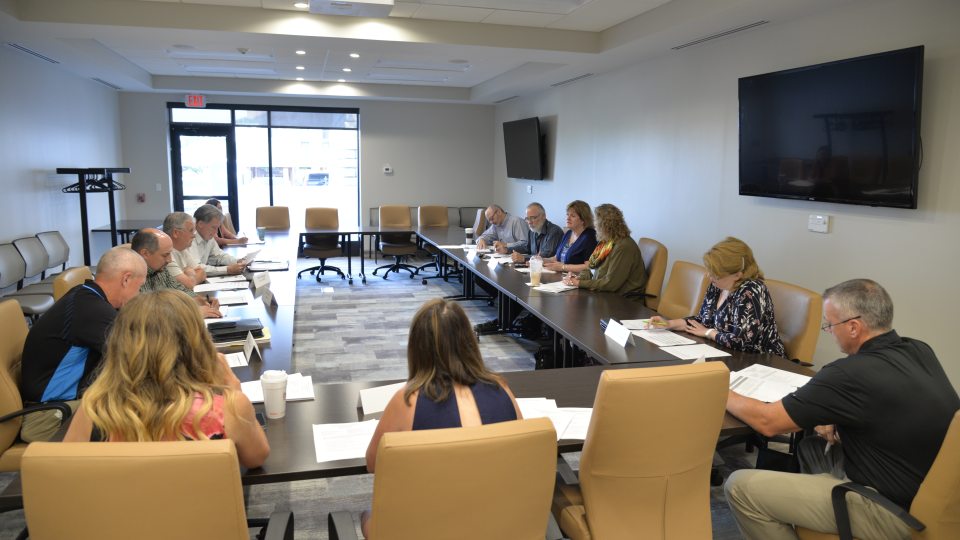 A group of people seated around a large U-shaped conference table in a modern meeting room. Participants are engaged in discussion, taking notes, and reviewing documents, with a projector and screen visible at the front of the room