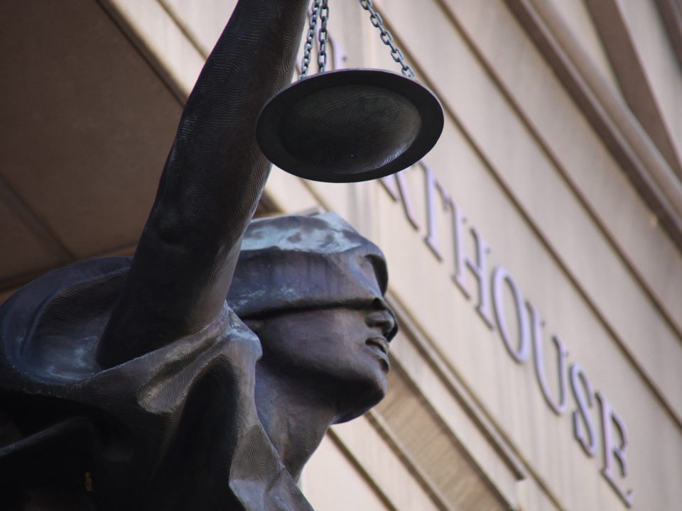 Close-up of a blindfolded statue of Lady Justice holding a balance scale, with the word 'Courthouse' engraved on the building in the background