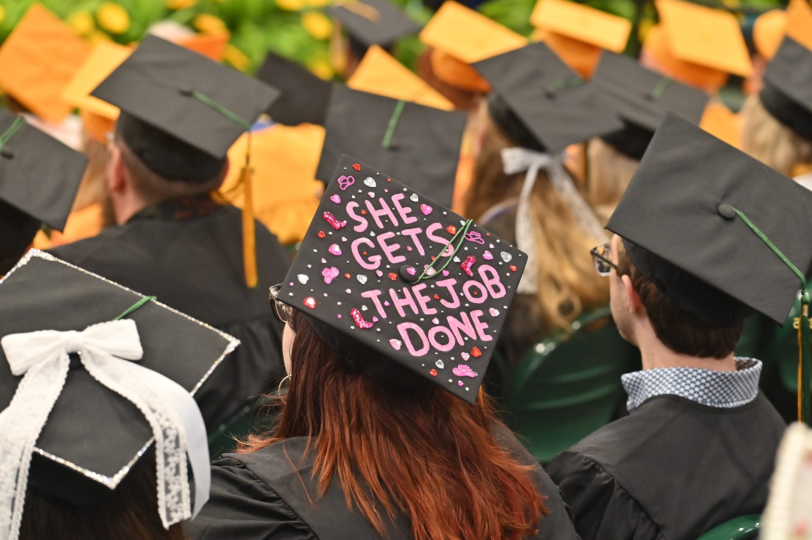 At commencement graduating students wear black and yellow hats; one hat reads "she gets the job done"