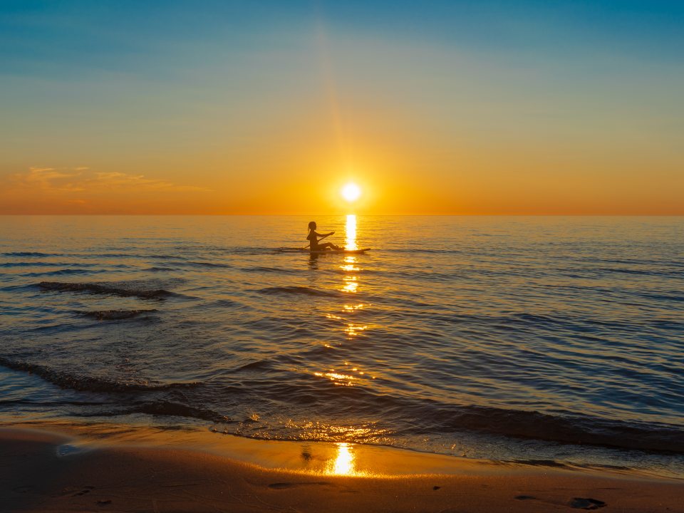 A person paddleboarding on Lake Ontario at sunset near Sandy Island in Oswego County, with the sun reflecting across the water toward the shore.