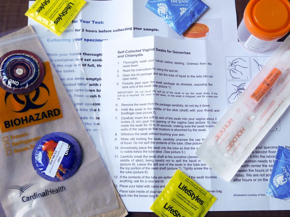 Image of an STI testing kit spread containing a biohazard bag, specimen cup, condoms, and instructions