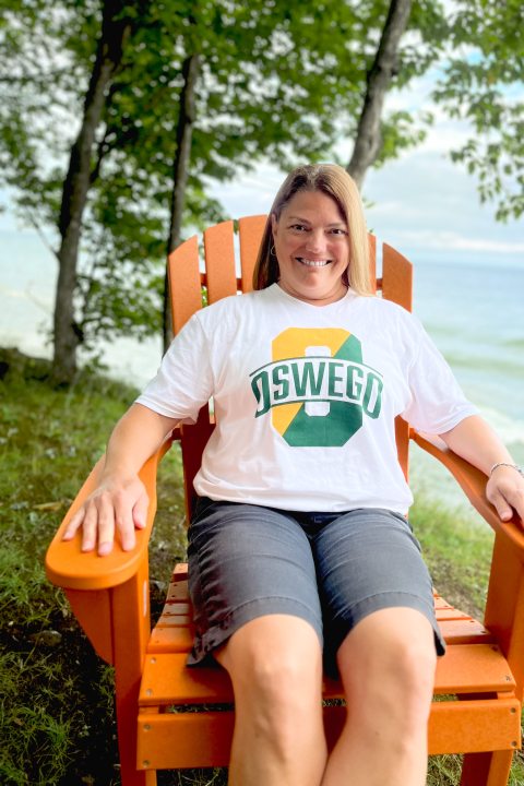 Angela sitting outdoors smiling for a portrait photo on a slightly cloudy day in the Summer. She is sitting on an Adirondack Chair with the view of Lake Ontario in the background, she also has a row of trees in view behind her..