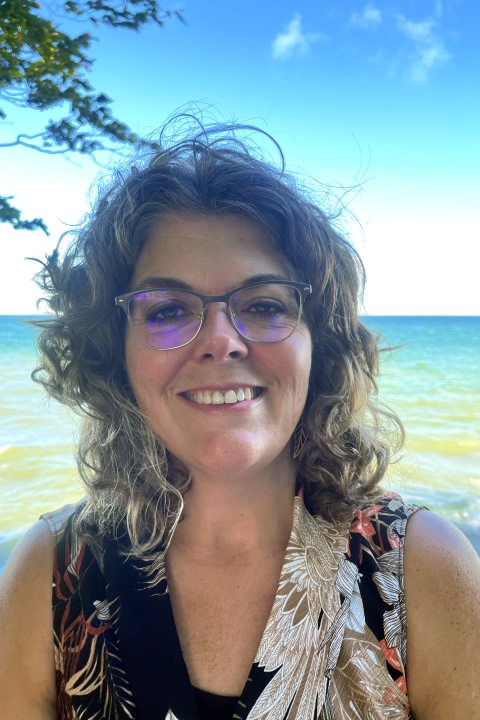Tricia outside on a sunny breezy day. She is standing in front of Lake Ontario surrounded by foliage