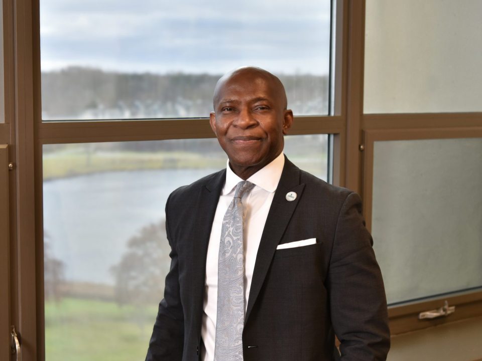 President Peter O. Nwosu standing in front of a window, overlooking the lagoon on the SUNY Oswego campus