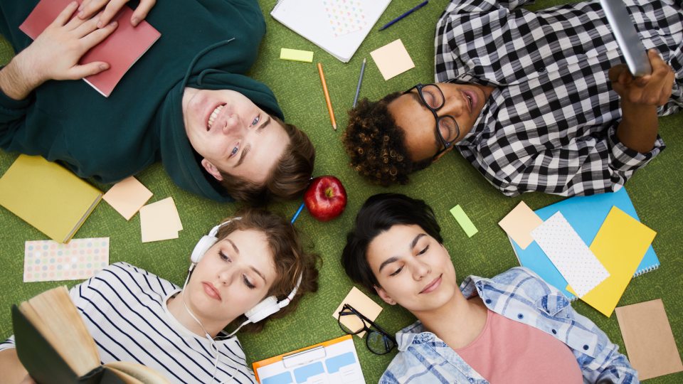 Birds eye view of students laying down on a lawn surrounded by notebooks and papers