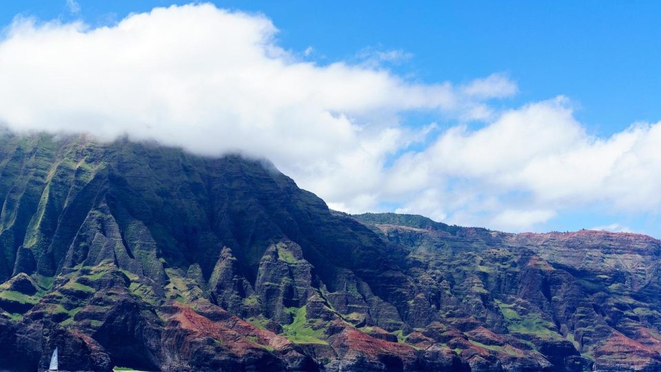 Steep green cliffs above blue ocean with a sailboat and low clouds