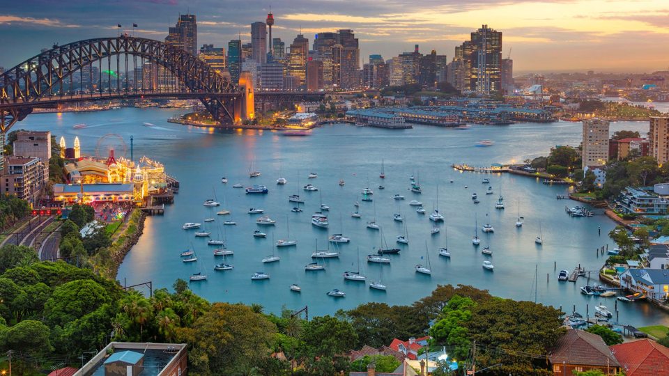 Sydney Harbour at dusk with the Harbour Bridge, illuminated city skyline, and sailboats on the water.