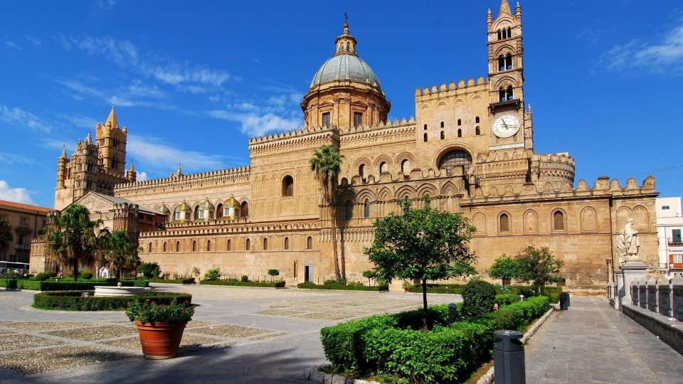 Palermo Cathedral with its iconic dome and towers overlooking a landscaped plaza under a bright blue sky.