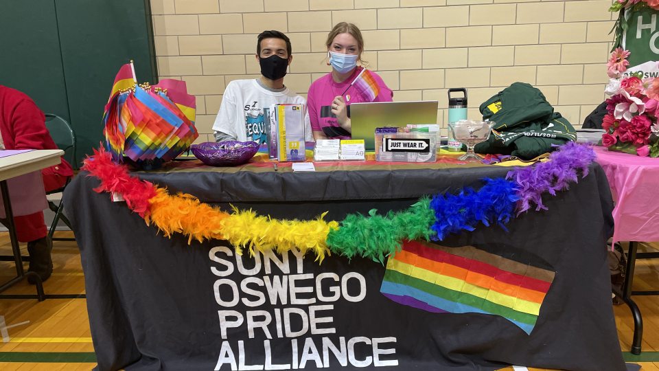 Students sitting behind a table with rainbow decorations and a black banner that reads SUNY Oswego Pride Alliance