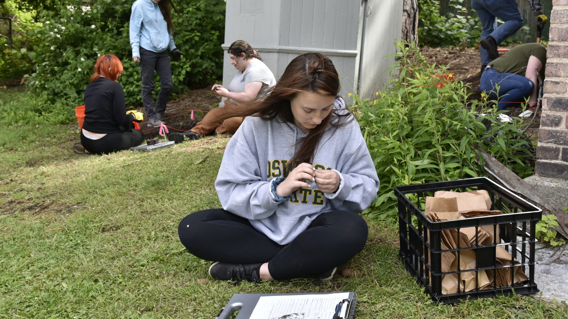 A group of students conduct fieldwork outdoors, with one sitting on the grass examining a sample while others dig and mark an area near a building, using tools and collection materials.
