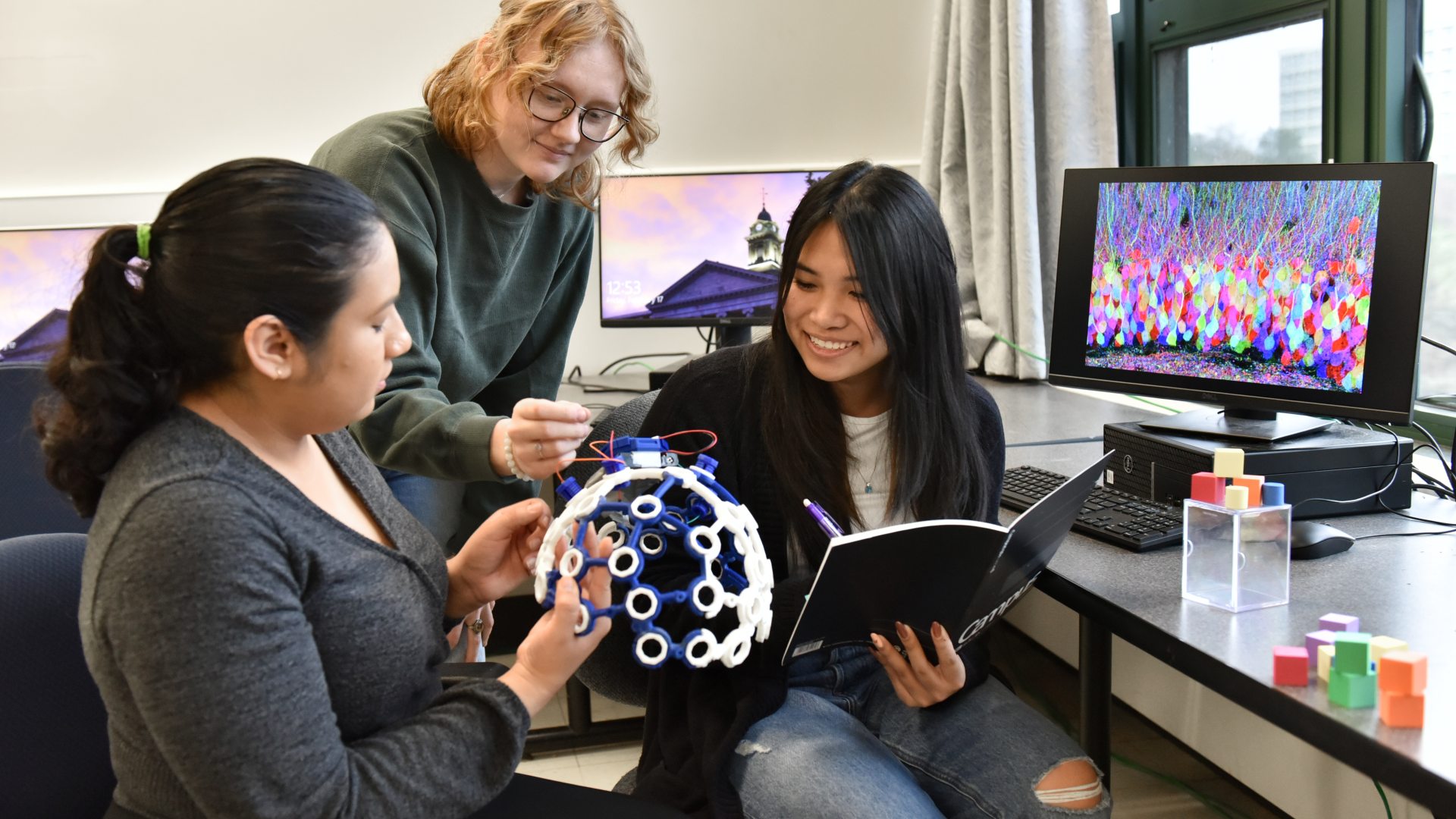 Three students collaborate in a lab, examining a geometric model while one takes notes, with computers displaying colorful visualizations in the background.