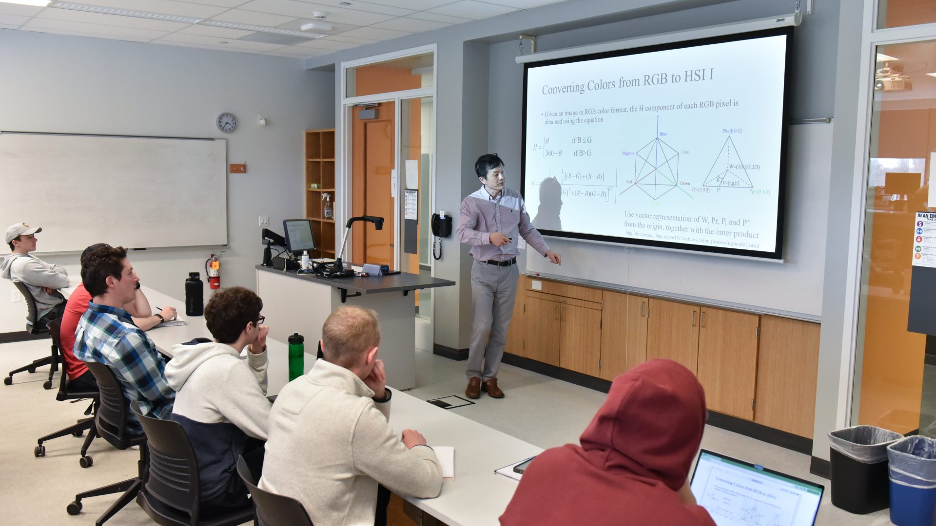 A professor teaches students in an engineering classroom, explaining color processing concepts on a projected screen.
