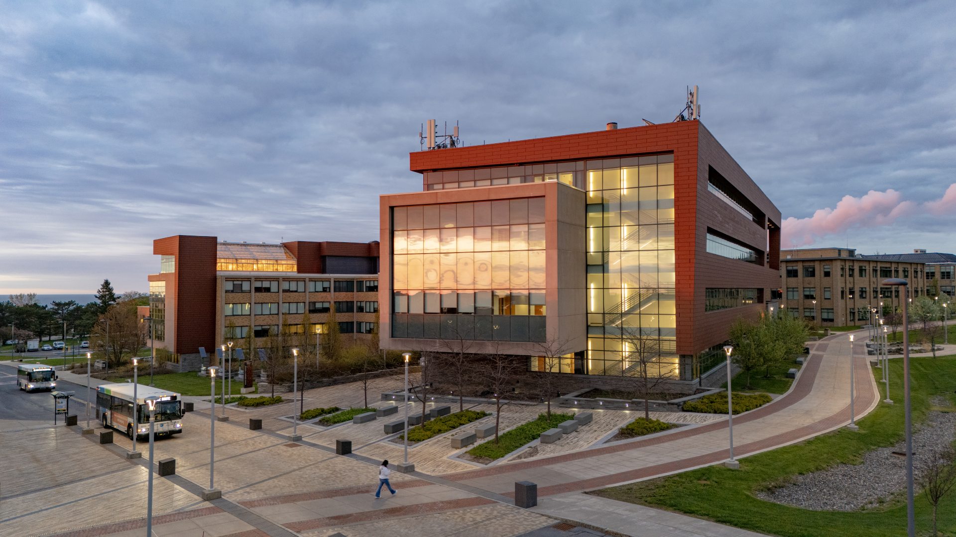 The Richard S. Shineman Center stands on SUNY Oswego’s campus, featuring modern architecture, solar panels, and a prominent vertical sign in a landscaped green space.