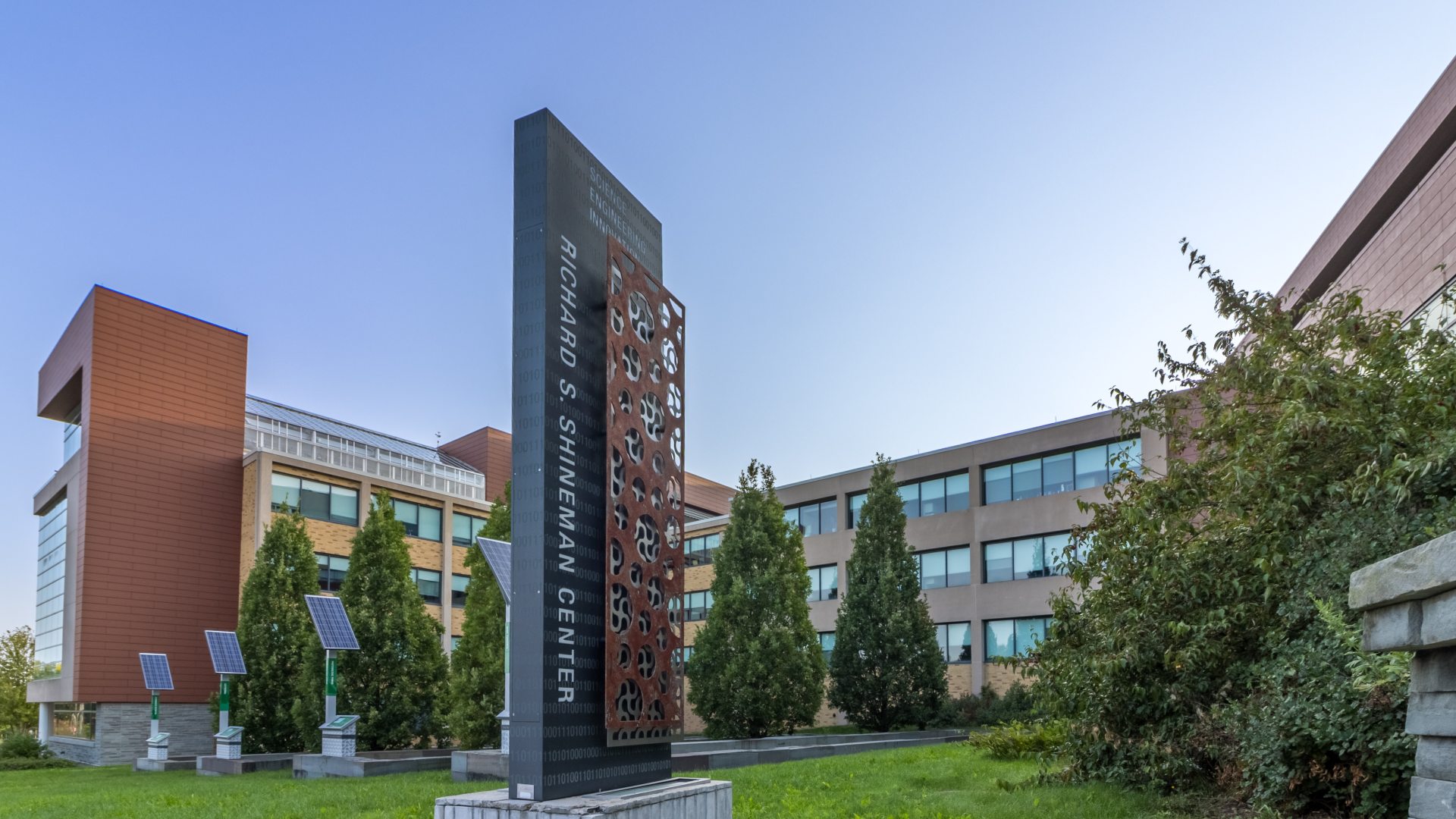 The Richard S. Shineman Center stands on SUNY Oswego’s campus, featuring modern architecture, solar panels, and a prominent vertical sign in a landscaped green space.