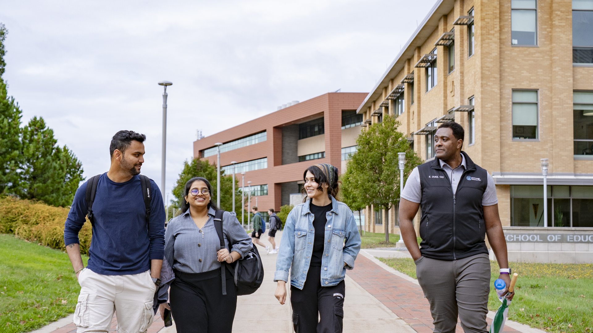 Four students walking and talking together along a campus path with academic buildings and trees in the background.
