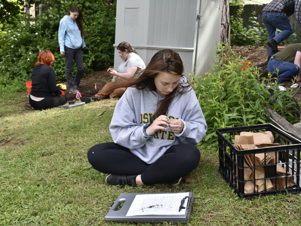 A group of students conduct fieldwork outdoors, with one sitting on the grass examining a sample while others dig and mark an area near a building, using tools and collection materials.