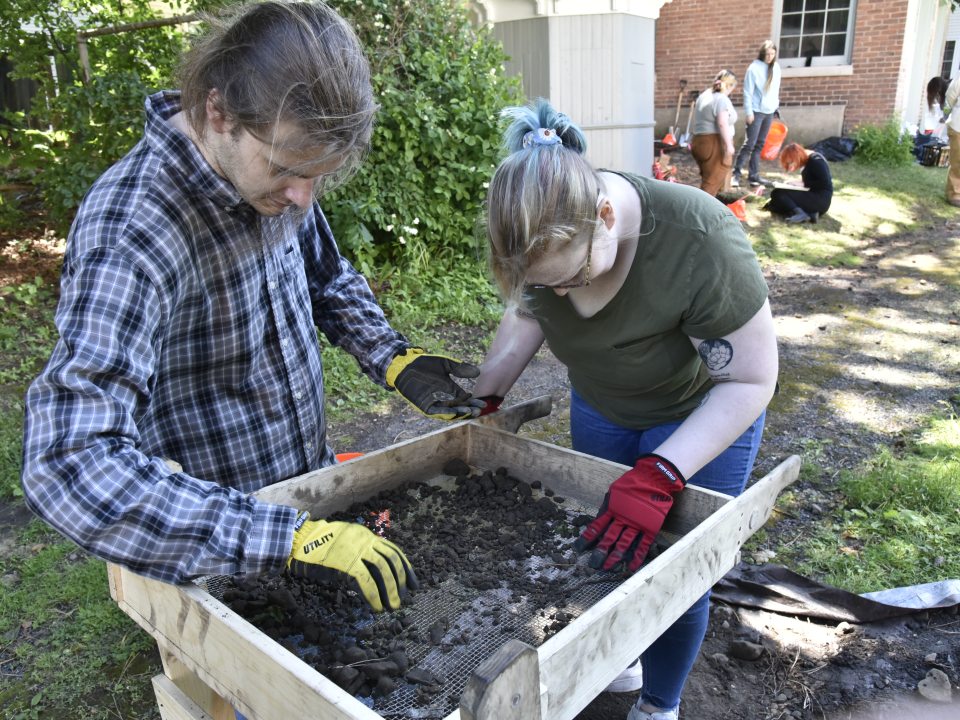 Students sift soil through a screen during an archaeological excavation as part of an anthropology field project.