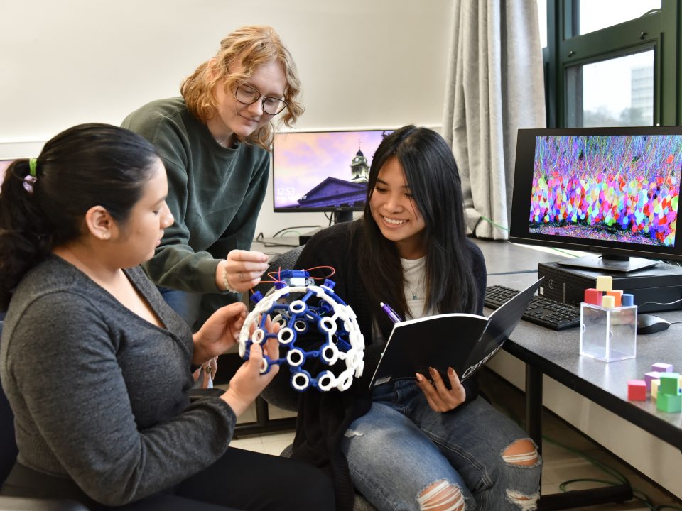 Three students collaborate in a lab, examining a geometric model while one takes notes, with computers displaying colorful visualizations in the background.