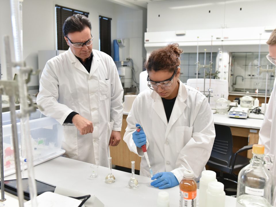 A professor works with students in a chemistry lab as one student uses a pipette during an experiment.