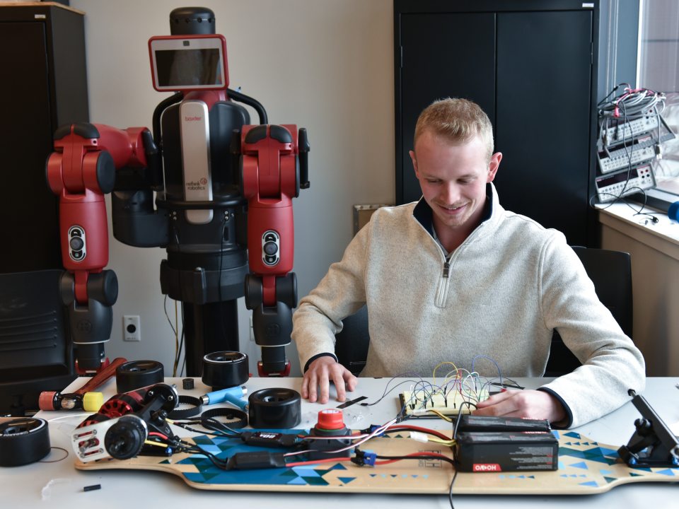 A student works on an electronics project with wires and components at a lab table, with a collaborative robot behind him in an engineering workspace.