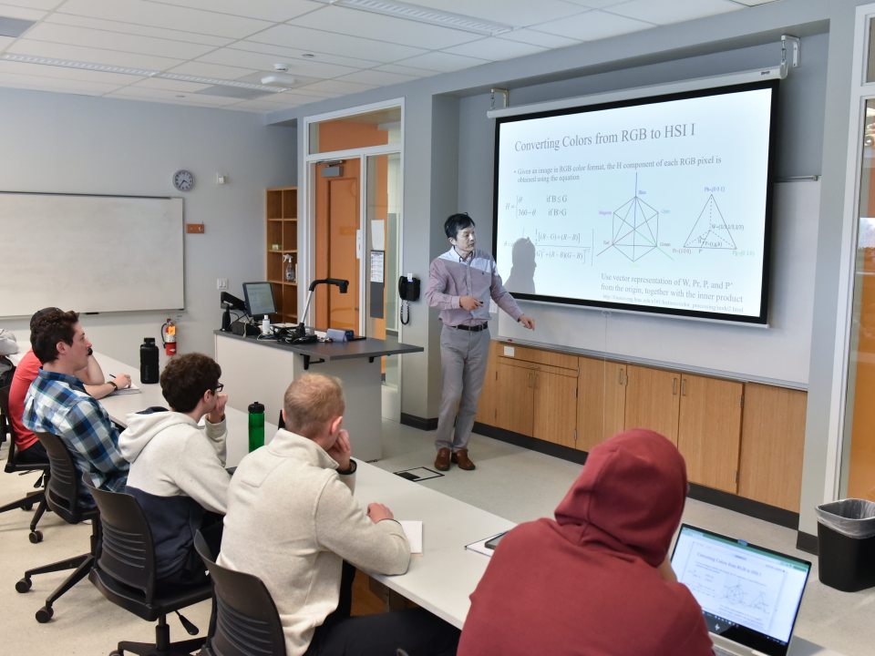 A professor teaches students in an engineering classroom, explaining color processing concepts on a projected screen.