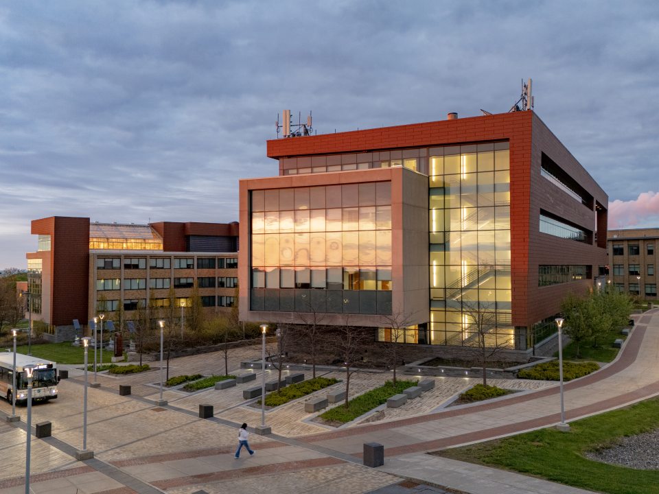 The Richard S. Shineman Center stands on SUNY Oswego’s campus, featuring modern architecture, solar panels, and a prominent vertical sign in a landscaped green space.
