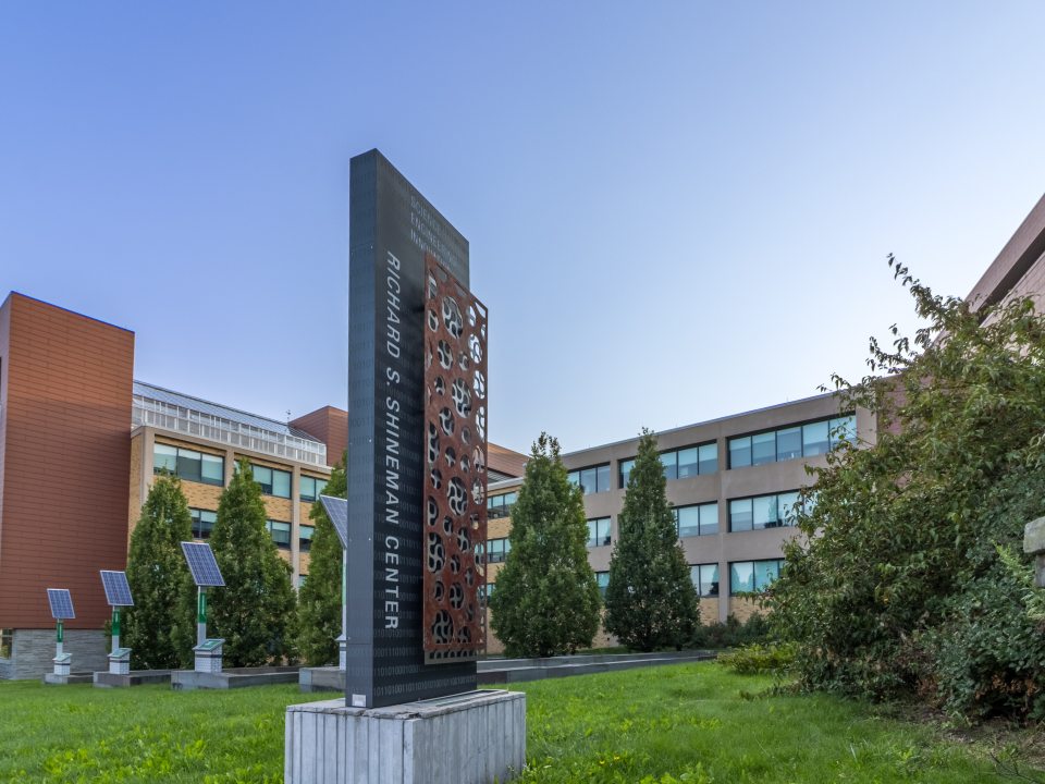 The Richard S. Shineman Center stands on SUNY Oswego’s campus, featuring modern architecture, solar panels, and a prominent vertical sign in a landscaped green space.