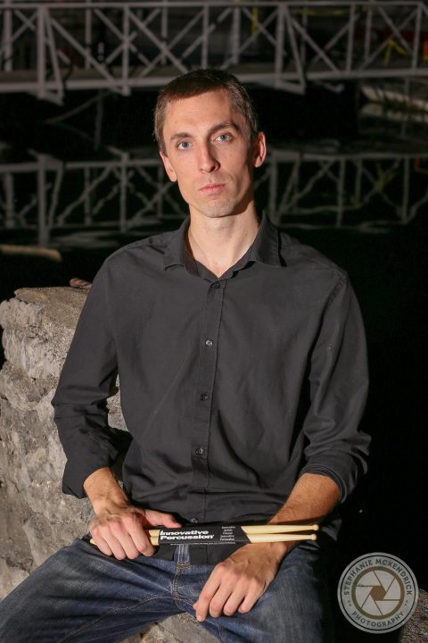 Nate Felty is sitting on some rocks holding drumsticks, wearing a black shirt and jeans