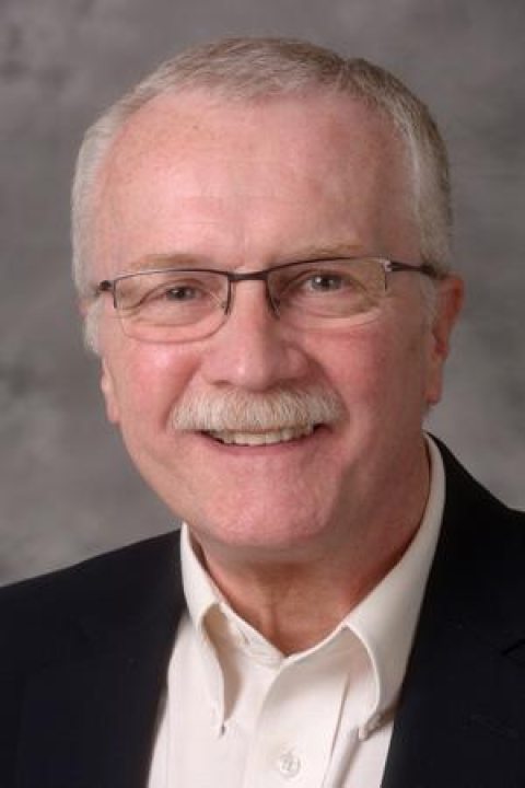 Robert Senko is wearing glasses and a black suit with a white shirt and smiling