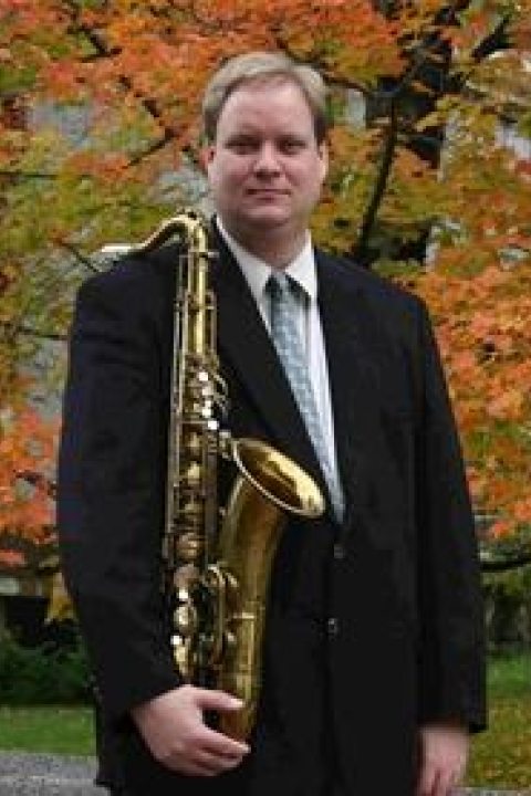 Trevor Jorgensen standing in front of a tree holding an instrument in a black suit, a white shirt and a green tie