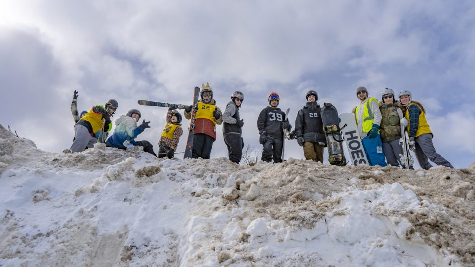 Skiers and snowboarders smile while standing atop a giant snow mound at a past Rail Jam