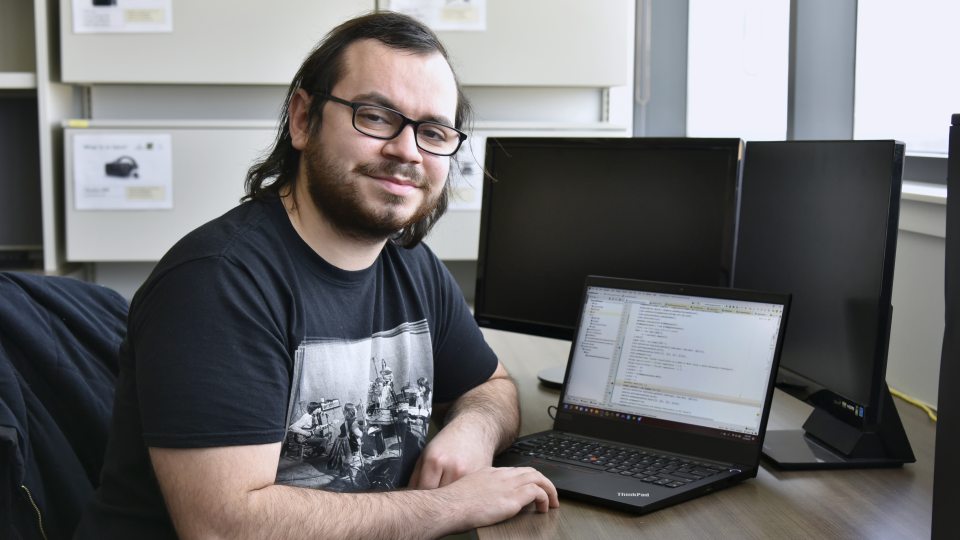 Computer science major Dominic Altamura sits in front of a computer, wearing a black t-shirt and black glasses. He has mid-length brown hair.