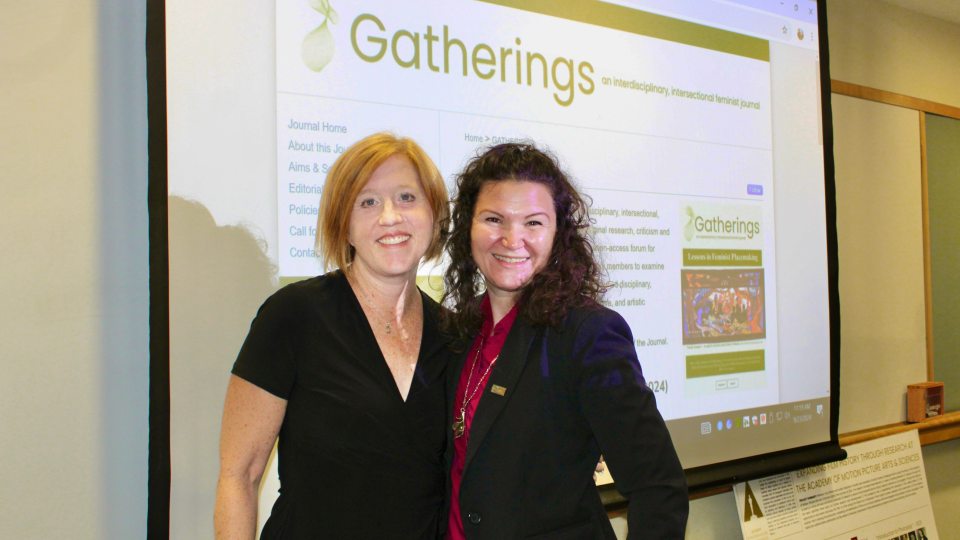 Cinema and Screen Studies professor Dr. Amy shore and English and Latin American studies studies professor Dr. Roberta Hurtado stand in front of the website for "Gatherings: An interdisciplinary and intersectional feminist journal."