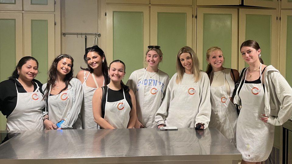 Students in the "Travel and Tourism" study abroad course pose for a picture at their cooking class held at Chefactory Cooking Academy in Florence, Italy. The group is all wearing white aprons with the company logo.