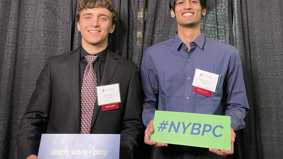SUNY Oswego students Paul Wehr and Harshit Gupta at the New York State Business Plan Competition, wearing red "finalist" badges.