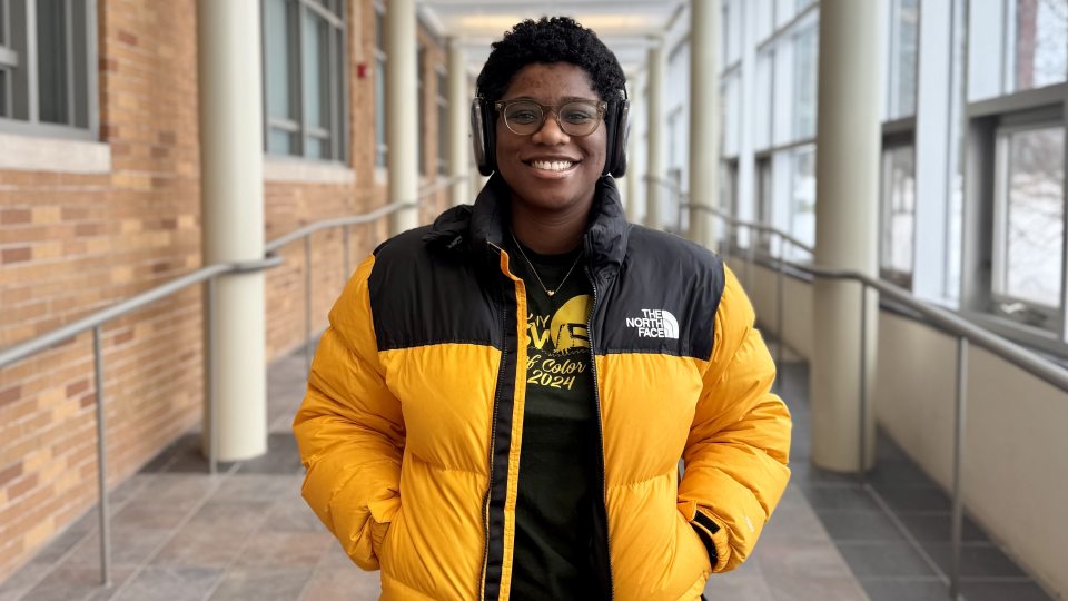 Oswego graduate student Keturah Hancock in Marano Campus Center. She wears a yellow coat and headphones.