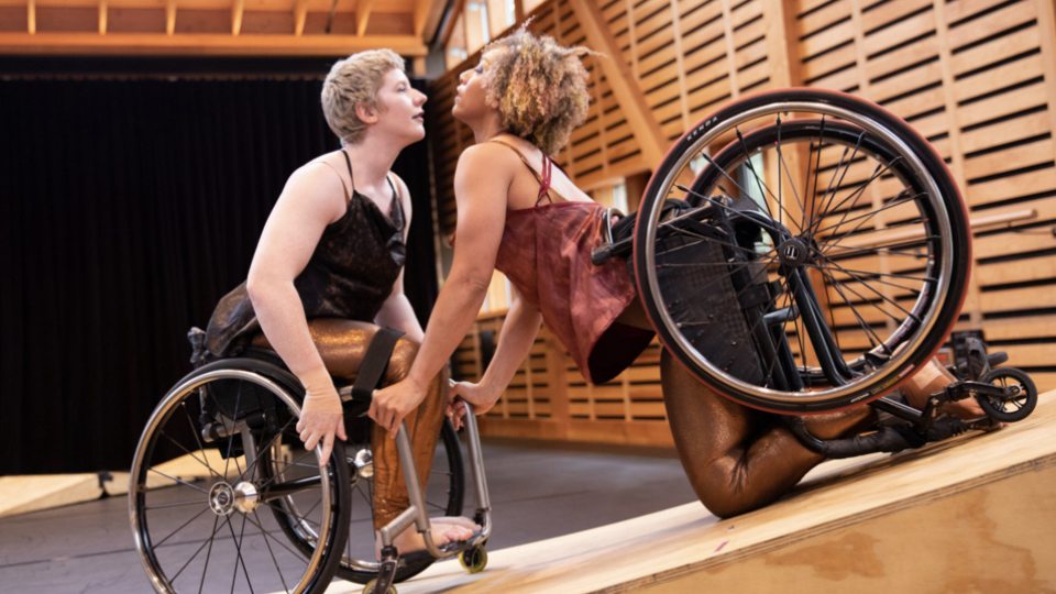 Both Alice and Laurel fly off the back end of the stage ramps looking at each other. Alice is a multiracial Black woman with short curly hair and coffee-colored skin; Laurel is a white dancer with pale skin and cropped blonde hair. They wear shimmery cost