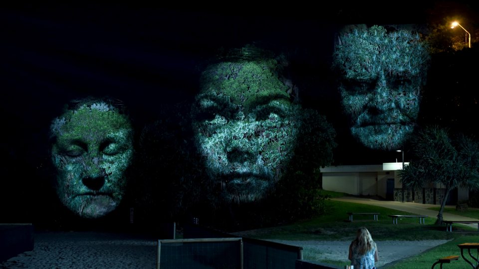Three projected faces appear on trees in a previous installation of Craig Walsh's Monuments