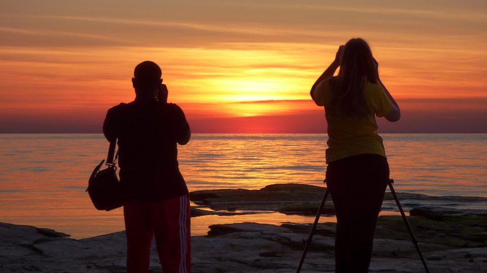 Two students take picture of an Oswego orange sunset over Lake Ontario