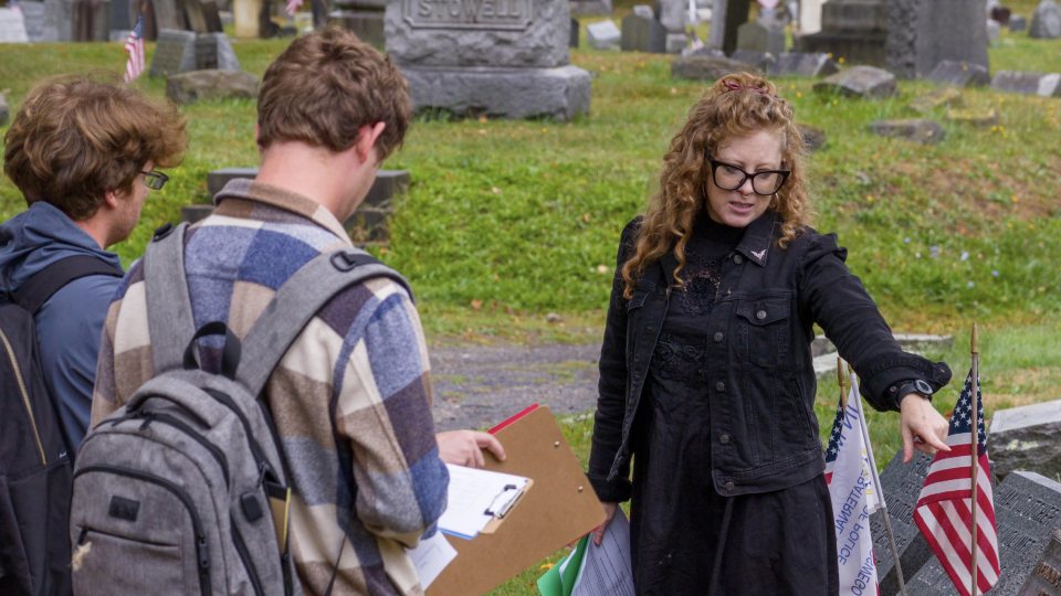 Oswego professor Candis Haak teaching her students in a local cemetery, pointing at a grave with an American flag.