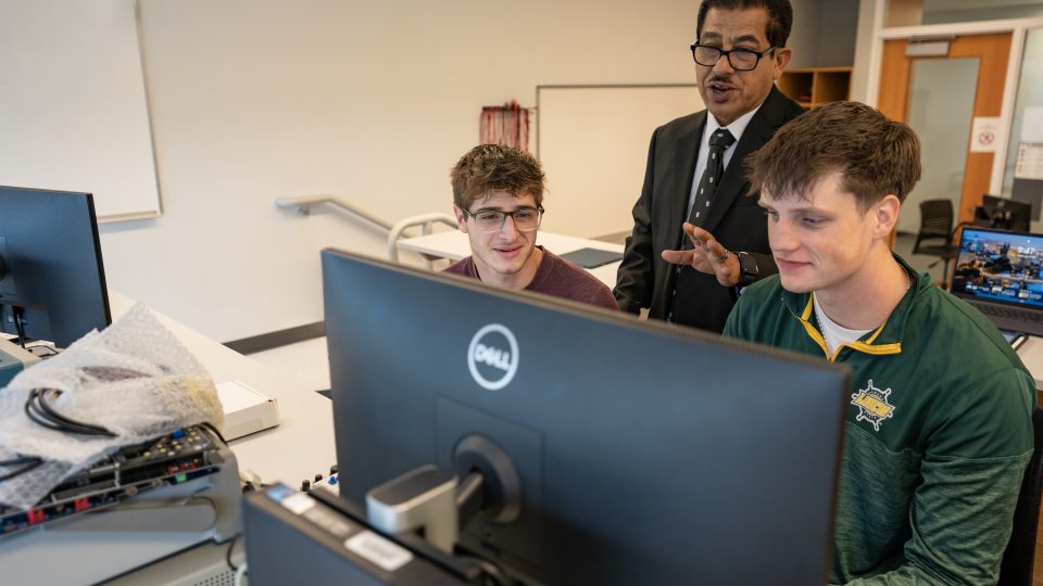 A faculty member stands behind two SUNY Oswego engineering students as they work together at a computer in an electrical and computer engineering lab.