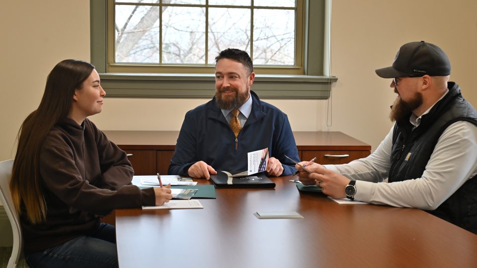 Three people sit around a conference table in a campus office, discussing materials during a meeting.
