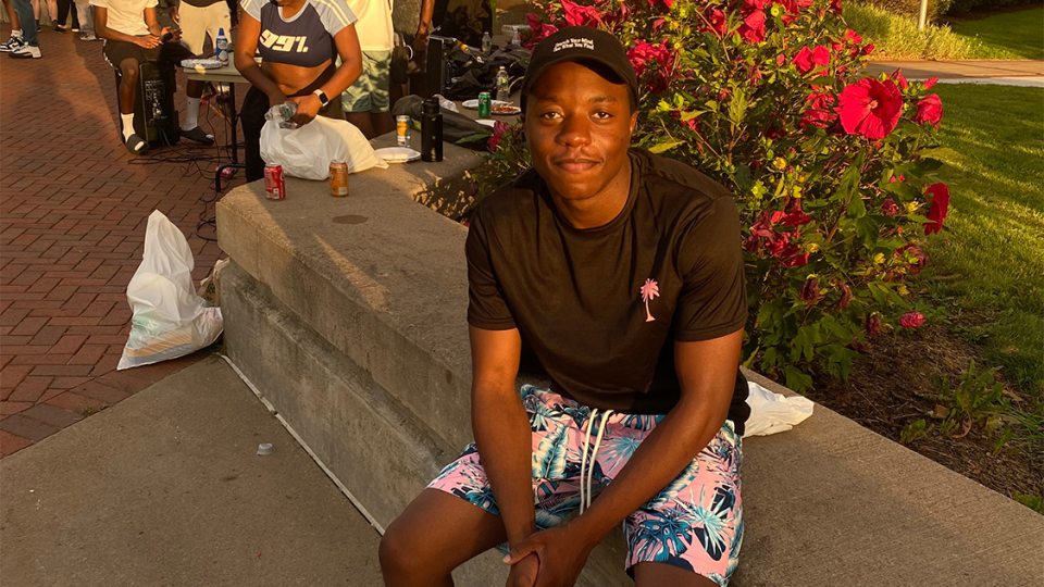 Antoine Okeke sits outside Hart Hall with the long line of students behind him waiting to try his menu for late night at Cooper Dining Hall. The sun is setting.