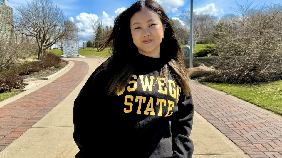 SUNY Oswego student Michelle Lau stands on campus in an black "Oswego State" sweatshirt.