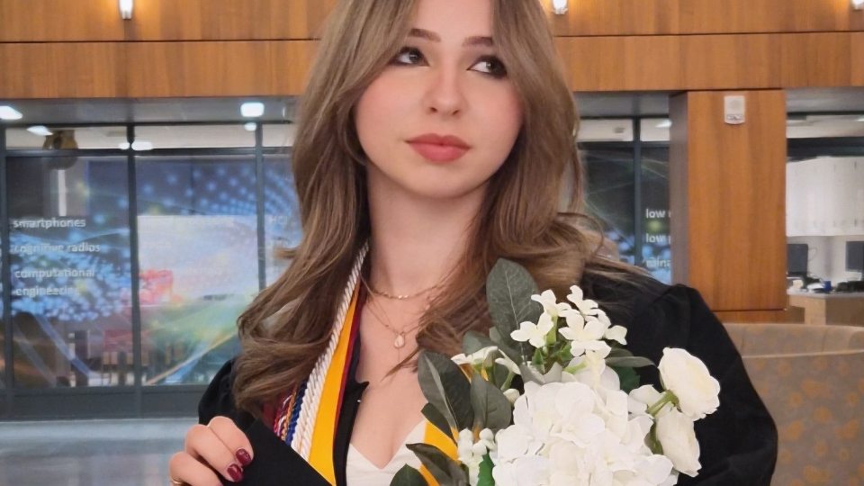 SUNY Oswego graduate Samantha Brown wearing a black cap and gown with honor cords and medals, holding a bouquet of white flowers while standing inside a modern campus building.