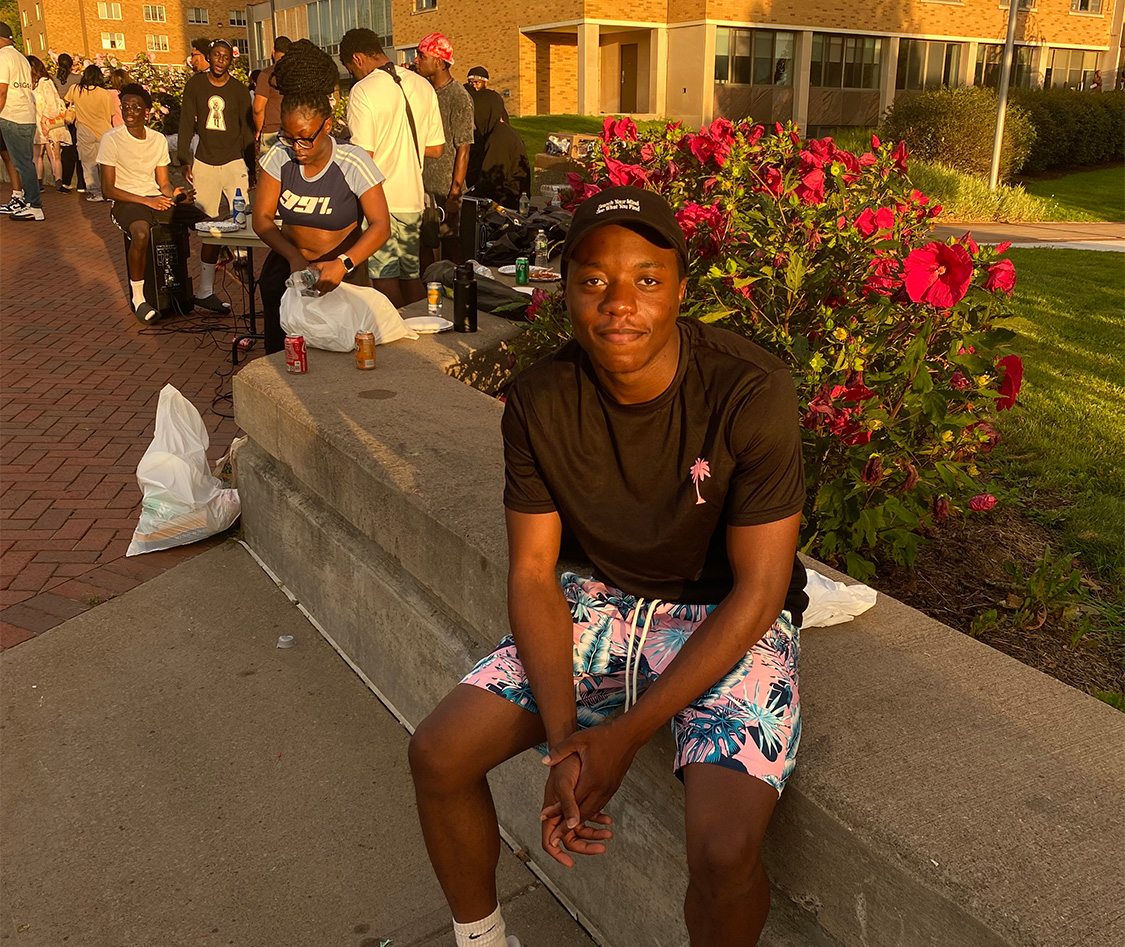 Antoine Okeke sits outside Hart Hall with the long line of students behind him waiting to try his menu for late night at Cooper Dining Hall. The sun is setting.