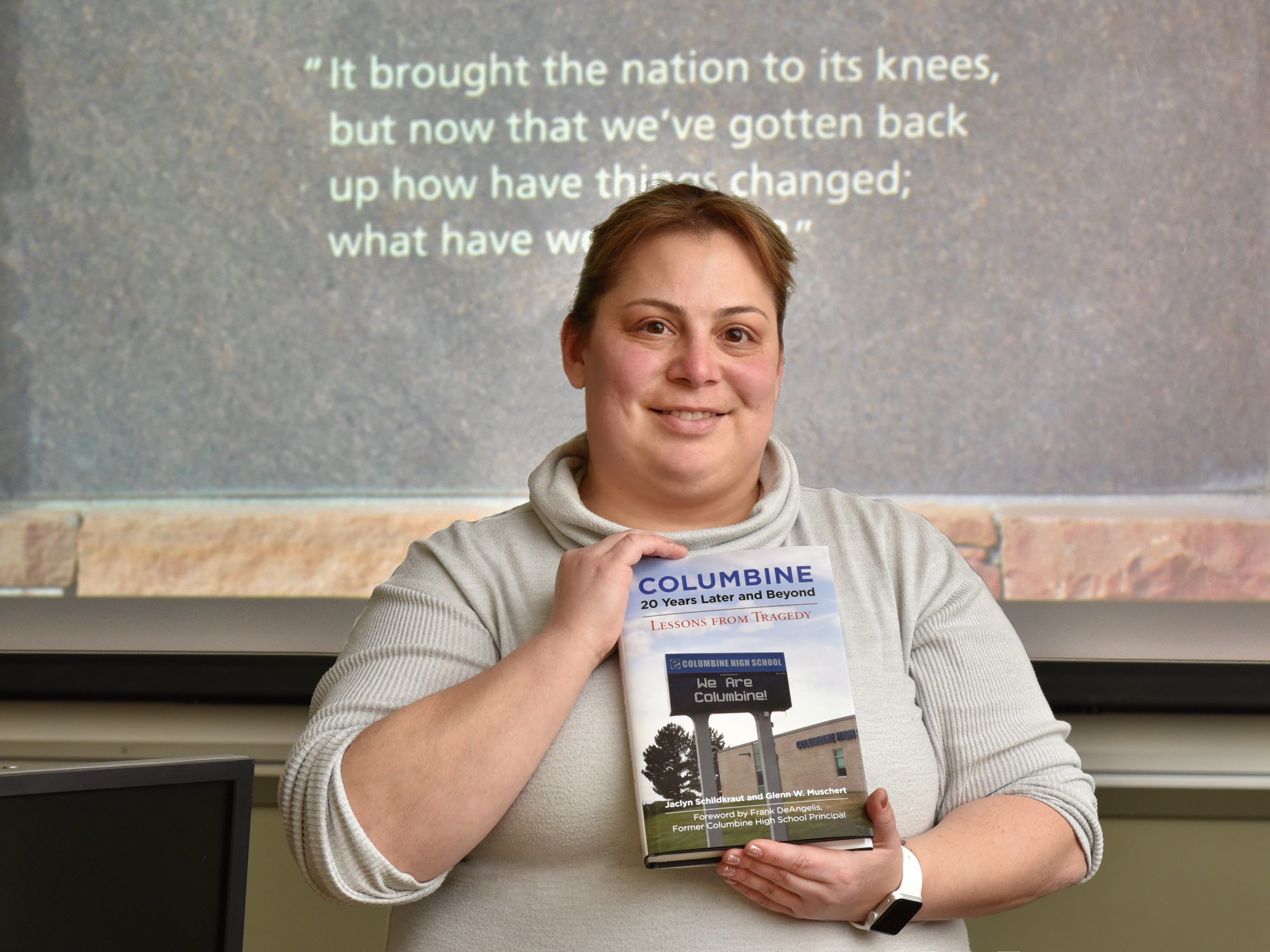 Jackie Schildkraut with book on 20th anniversary of Columbine, with quote from healing wall on screen behind her