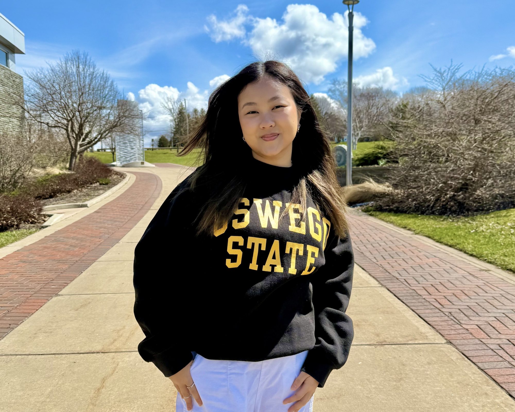 SUNY Oswego student Michelle Lau stands on campus in an black "Oswego State" sweatshirt.
