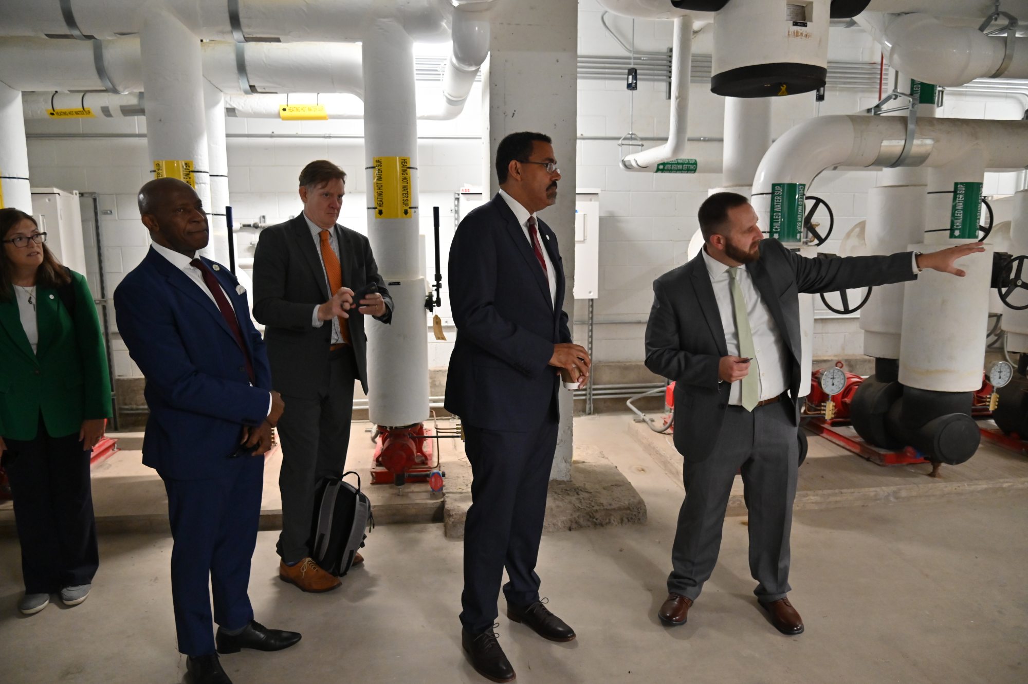 Five people tour a campus mechanical room as a staff member gestures toward large pipes and building equipment.