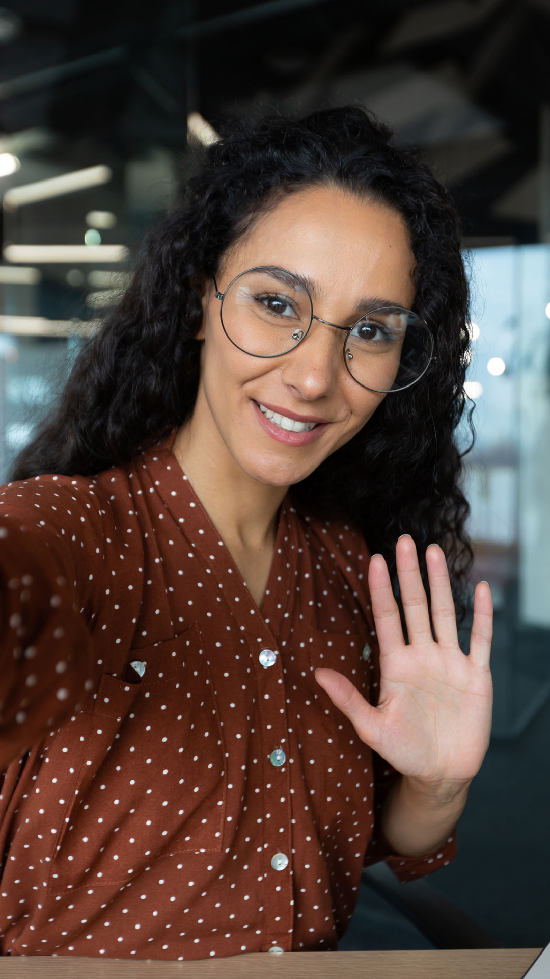 Young woman with curly dark hair and glasses waves and greets visitors. 